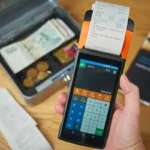 Mesin kasir mencetak struk nota kontan dengan uang tunai dan koin terlihat di samping meja transaksi